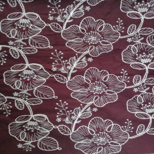 Large stylized two tone floral motifs are outlined with thick, rope-like embroidery, giving it a 3D effect on a chocolate colour linen look fabric base.