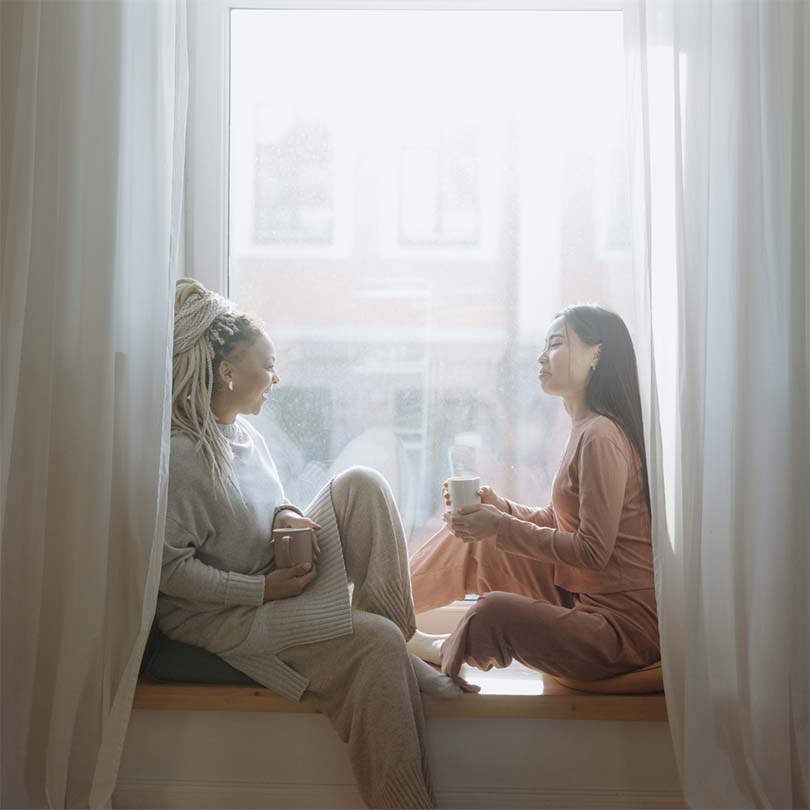 2 chatting girls sitting in the morning sun in a window seat with coffee, dressed in lounge wear.