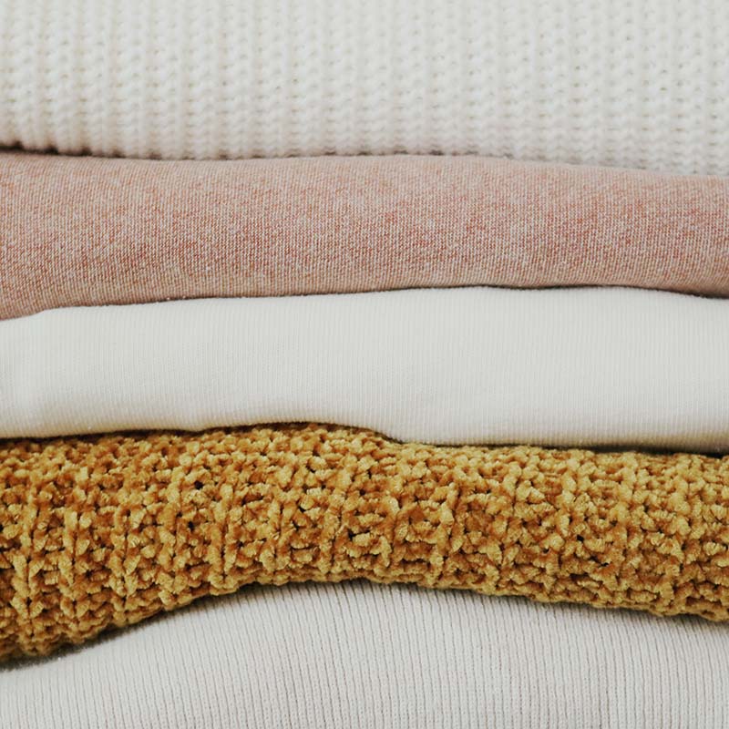 A stack of folded textured knitted fabrics in pastel and neutral colours.